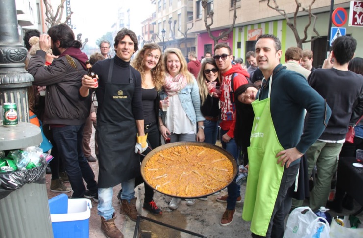 スペイン、バレンシアのスペイン語学校costa de valenciaのアクティビティで屋外で大きなパエリアと一緒に写真をとる生徒と、背景にたくさんの人たちが写っている