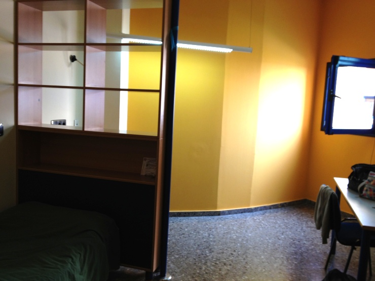 黄色い壁と緑のベッドカバーが敷かれたベッドがある学生寮の部屋の一角